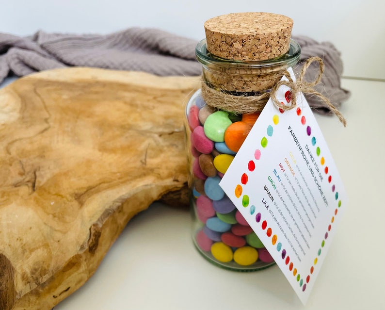 May include: A glass jar filled with colorful candy, tied with twine and a tag that reads "Danke f&uuml;r die farbenfrohe Zeit und sch&ouml;ne Zeit!" with a color key for orange, red, blue, green, brown, and purple.