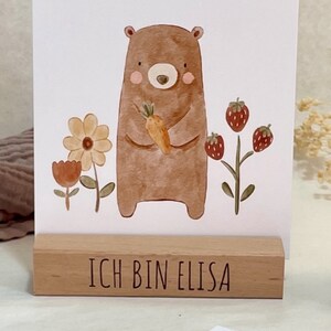 K&ouml;nnte beinhalten: Ein brauner B&auml;r, der eine Karotte h&auml;lt, steht vor einem wei&szlig;en Hintergrund mit dem Text "Ich bin stark" oben. Der B&auml;r ist von Blumen und Erdbeeren umgeben. Ein Holzblock mit dem Text "Ich bin Elisa" befindet sich unter dem B&auml;ren.