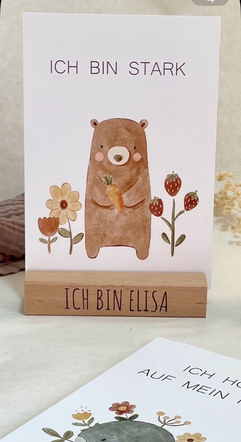 K&ouml;nnte beinhalten: Ein brauner B&auml;r, der eine Karotte h&auml;lt, steht vor einem wei&szlig;en Hintergrund mit dem Text "Ich bin stark" oben. Der B&auml;r ist von Blumen und Erdbeeren umgeben. Ein Holzblock mit dem Text "Ich bin Elisa" befindet sich unter dem B&auml;ren.