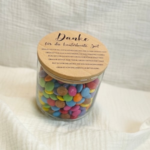 May include: A clear glass jar filled with colorful candies, topped with a wooden lid. The lid has engraved text in German. The jar is sitting on a white, textured surface.