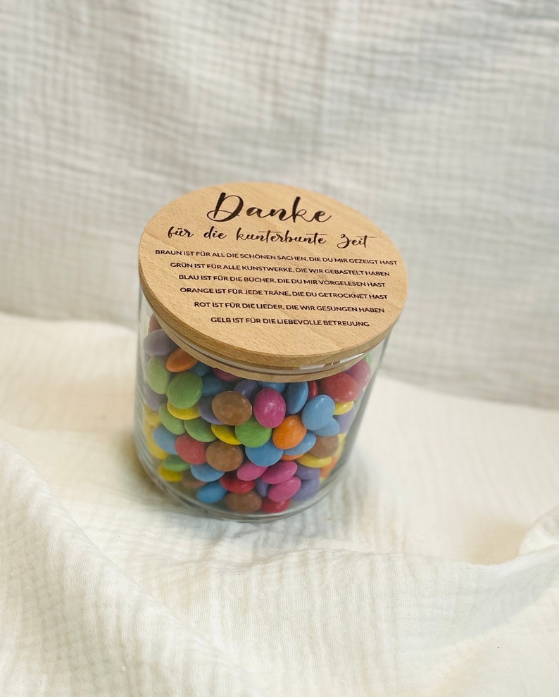 May include: A clear glass jar filled with colorful candies, topped with a wooden lid. The lid has engraved text in German. The jar is sitting on a white, textured surface.