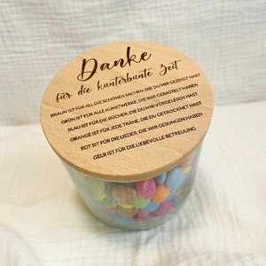 May include: A clear glass jar filled with colorful candies, topped with a wooden lid. The lid is engraved with the German phrase "Danke f&uuml;r die kunterbunte Zeit" and other text. The jar sits on a white, textured surface.