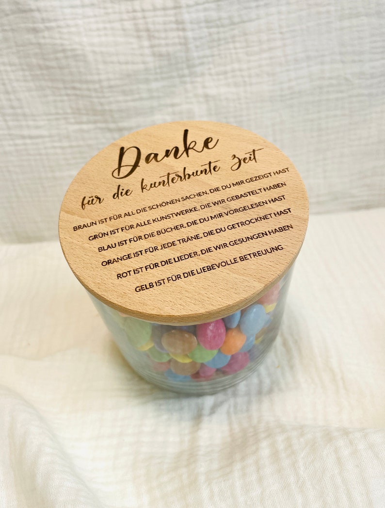 May include: A clear glass jar filled with colorful candies, topped with a wooden lid. The lid is engraved with the German phrase "Danke f&uuml;r die kunterbunte Zeit" and other text. The jar sits on a white, textured surface.