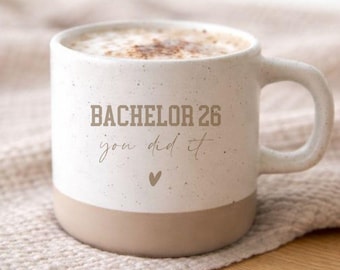 May include: A speckled white ceramic mug with a smooth tan base. The front features the text BACHELOR 26 in a bold block font, followed by the script text you did it and a small heart icon. The mug is filled with a frothy beverage.