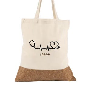 Könnte beinhalten: Eine cremefarbene Stofftasche mit zwei Henkeln, verziert mit einer schwarzen Grafik eines Stethoskops, das eine Herzschlaglinie und ein Herz bildet. Unter der Grafik ist der Name SARAH in schwarzen Großbuchstaben gedruckt. Der Boden ist aus Kork.