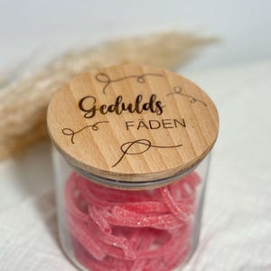 Könnte beinhalten: Ein klares Glas mit rosa Gummibonbons, bedeckt mit einem Holzdeckel. Der Deckel ist mit den Worten "Gedulds Fäden" in einer dekorativen Schrift graviert. Das Glas steht vor einem weichen, verschwommenen Hintergrund.