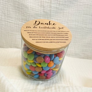 May include: A clear glass jar filled with colorful candies, topped with a wooden lid. The lid has the German text "Danke f&uuml;r die kunterbunte Zeit" and other text. The jar is filled with various colors of candies.
