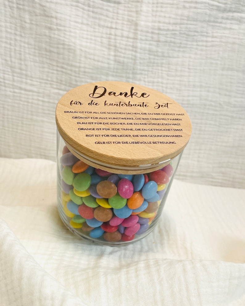 May include: A clear glass jar filled with colorful candies, topped with a wooden lid. The lid has the German text "Danke f&uuml;r die kunterbunte Zeit" and other text. The jar is filled with various colors of candies.