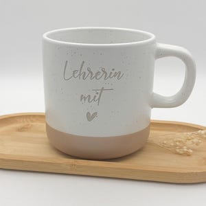 Könnte beinhalten: Weiße Keramiktasse mit gesprenkeltem Finish und einem hellbraunen Band um den Boden. Die Tasse hat den Text "Lehrerin mit" und ein kleines Herz auf der Vorderseite.
