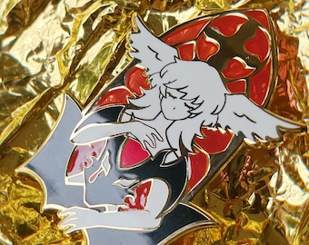 May include: A black and red enamel pin featuring a white angel with wings, holding a heart in its hands. The angel is surrounded by a black and red design.