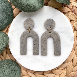May include: A pair of gray speckled clay earrings with a gold accent. The earrings are shaped like an arch with a round disc at the top.