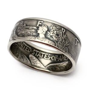 May include: A silver ring with detailed engravings. The ring features the words "UNITED STATES OF AMERICA" and images of a woman and foliage. The ring has a wide band and a vintage aesthetic.