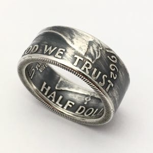 Könnte beinhalten: Ein silberner Ring aus einer Half-Dollar-Münze von 1962. Der Ring trägt die Inschriften "IN GOD WE TRUST" und "HALF DOLLAR". Der Ring hat ein detailliertes, strukturiertes Design. Ein einzigartiges Schmuckstück.