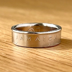 May include: A silver ring with the word "JAMAICA" engraved on the band. The ring has a textured edge and is set against a wooden surface. The ring is a piece of jewelry.