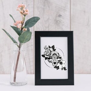May include: Black and white illustration of a cat skull with flowers. The skull is surrounded by a circle and the flowers are in bloom. The illustration is framed in a black frame.
