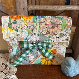 May include: A patchwork clutch bag with a vibrant mix of floral and illustrated patterns. It's fastened with a green and white checked bow. A light blue ceramic bird is placed on the side.