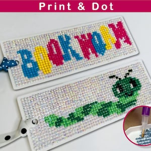 Op de afbeelding: Twee witte bladwijzersets met kleurrijke ruitvormige kralen. Eén bladwijzer heeft het woord "BOOKWORM" in kleurrijke kralen. De andere bladwijzer heeft een groene rups met een glimlachend gezicht. De afbeelding toont ook een close-up van een dienblad met blauwe kralen en een gereedschap om de kralen te plaatsen.