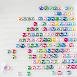 May include: Rows of iridescent, colorful rhinestones arranged in a grid pattern on a white surface. The rhinestones are various shades of blue, green, red, pink, and white.