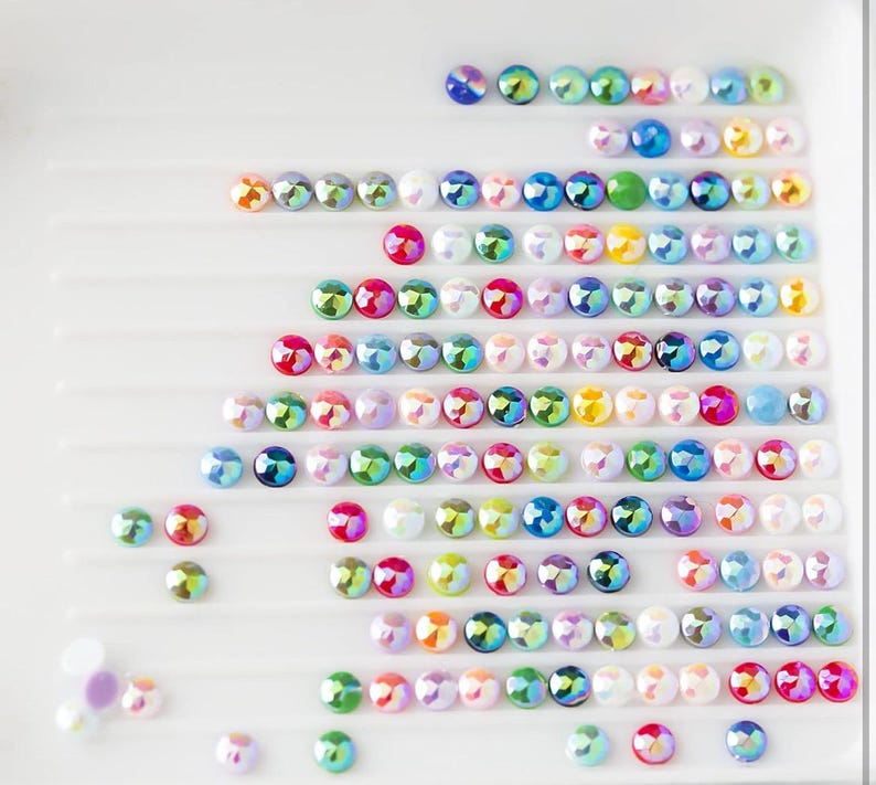 May include: Rows of iridescent, colorful rhinestones arranged in a grid pattern on a white surface. The rhinestones are various shades of blue, green, red, pink, and white.