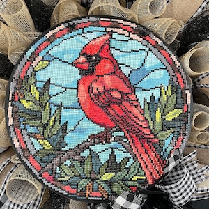 May include: A red cardinal bird with black eyes and a yellow beak is perched on a branch in a stained glass style design. The background is a blue sky with green trees. The design is on a round, black, wooden plaque.