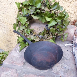 Può includere: Una padella di ghisa nera con un manico lungo si trova su una superficie di pietra. Una ghirlanda di foglie verdi con bacche scure è dietro la padella.