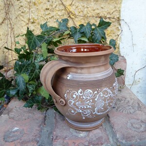 Könnte beinhalten: Ein brauner Keramik Krug mit einem weißen Blumenmuster. Der Krug hat einen Henkel und eine breite Öffnung.
