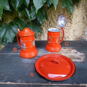 Könnte beinhalten: Drei rote Emaille-Becher mit schwarzem Rand. Die Becher haben ein Blumenmuster und einen Henkel. Ein Becher hat einen Deckel.
