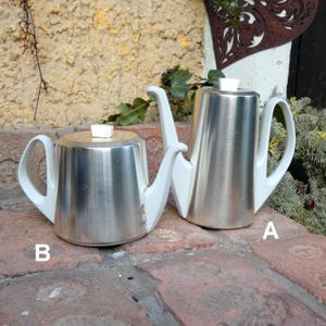 Könnte beinhalten: Zwei silberne Teekannen mit weißen Griffen und Deckeln. Die Teekanne links ist mit "B" und die Teekanne rechts mit "A" gekennzeichnet.