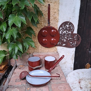 Könnte beinhalten: Eine Sammlung von Küchenutensilien in Rot und Weiß. Dazu gehören eine Pfanne mit vier runden Formen, ein kleiner Eimer, zwei Becher und eine Pfanne mit langem Griff. Ein dekoratives Metallherz ist ebenfalls vorhanden.