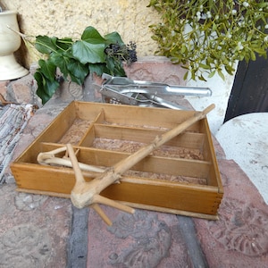 May include: A wooden organizer with multiple compartments, a wooden tool, and a metal nutcracker. Green leaves and berries are arranged around the organizer. The wooden organizer is a light brown color.