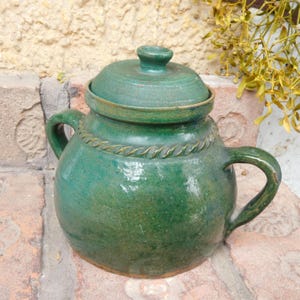 Pode incluir: Um pote de cerâmica verde com tampa e duas alças. O pote tem formato redondo e superfície texturizada. A tampa tem um pequeno botão no topo. Um design decorativo em forma de corda envolve o pote. O pote está sobre uma superfície de tijolo.