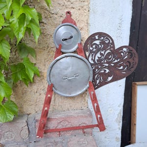 Puede incluir: Adorno de pared rústico con dos tapas de olla metálicas sujetas por una estructura de madera roja en forma de escalera. Un corazón decorativo de metal oxidado con un diseño ornamental también está presente. Fondo texturizado, claro, con hojas verdes.
