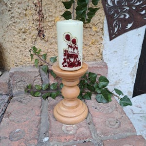 May include: A white pillar candle with a red and gold design of a family on a wooden candle holder.