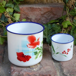 Op de afbeelding: Twee witte emaille bekers met blauwe randen. De grotere beker heeft een bloemenmotief met rode klaprozen en groene bladeren op een wit en blauw geruit patroon. De kleinere beker heeft een eenvoudig ontwerp met groene bladeren en rode bessen op een witte achtergrond.