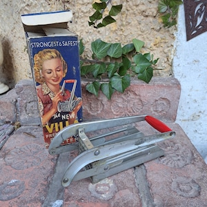 Op de afbeelding: Vintage metalen frietjes-snijder met een rode handgreep. De snijder ligt op een bakstenen oppervlak. De doos voor de snijder is op de achtergrond. De doos is blauw en wit en heeft een afbeelding van een vrouw die de snijder gebruikt. Op de doos staat "The New Villa French Fryer".
