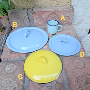 May include: A light blue enamel mug with a dark blue rim sits next to three enamel lids in light blue and yellow. The lids have a handle on top. The items are on a brick surface.