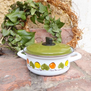 Può includere: Una pentola di smalto bianco con un coperchio verde e un pomello nero. La pentola ha un design floreale con accenti arancioni e gialli.
