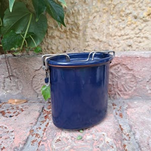 Op de afbeelding: Een marineblauwe emaille pot met een zilveren metalen sluiting en rand. De cilindrische pot heeft een afgeronde bodem en staat op een bakstenen oppervlak met groen op de achtergrond.