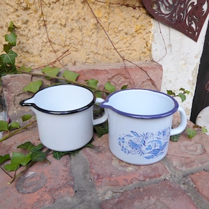 Może przedstawiać: Dwa białe emaliowane kubki z czarnymi obwódkami. Jeden kubek ma niebieski i biały wzór kwiatowy.