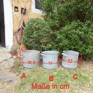 Op de afbeelding: Drie gegalvaniseerde metalen plantenbakken met handvatten, gelabeld A, B en C. Plantenbak A heeft een diameter van 36,5 cm, terwijl plantenbakken B en C een diameter van 32 cm hebben. De plantenbakken staan op een bed van groen gras.