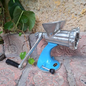 Op de afbeelding: Een vintage handmatige vleesmolen met een zilverkleurige metalen behuizing en een helderblauwe basis. De molen heeft een handslinger met een zwarte handgreep en een invoerbak. De basis is gemarkeerd met het nummer "805".