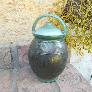 Op de afbeelding: Een handgemaakte keramische pot met een deksel en handvat. De pot heeft een donkergroene, getextureerde body met bloemen- en geometrische patronen. Het deksel en het handvat zijn een lichtere tint groen. De pot is ongeveer 25 cm hoog.