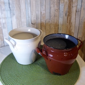 May include: Two vintage enamelware crocks, one white and one red, with a dark gray liquid inside the red crock. The crocks are sitting on a green circular placemat.