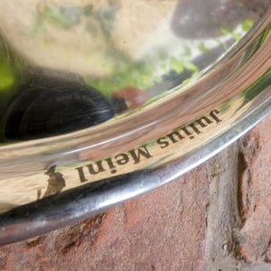 Pode incluir: Uma bandeja de serviço circular de metal prateado com uma superfície polida e reflexiva. A borda da bandeja apresenta o texto gravado Julius Meinl em uma fonte serifada escura. A superfície metálica mostra leves arranhões e reflexos.