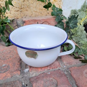 Puede incluir: Tazón de esmalte blanco con borde azul y una pequeña pegatina en el lateral. El tazón tiene una sola asa.