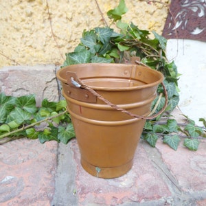 Op de afbeelding: Een koperen emmer met een draadhendel. De emmer staat op een bakstenen oppervlak met groene klimop planten eromheen.