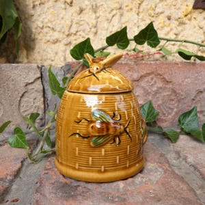 Könnte beinhalten: Beigefarbener Keramik-Honigtopf in Bienenkorbform mit Deckel. Er hat eine strukturierte Oberfläche und Bienenmotive an der Seite und am Deckel. Der Hintergrund zeigt Ziegelmauerwerk und grünes Laub.