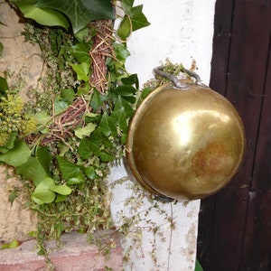 Op de afbeelding: Een messing kom met een handvat hangt aan een krans van groene klimop. De kom is glanzend en heeft een licht verweerde patina.