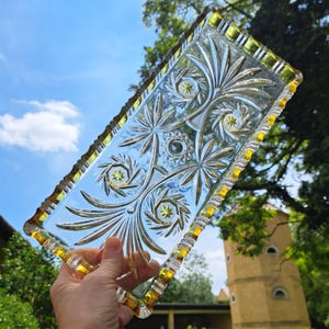 May include: A rectangular, clear glass tray with intricate floral and starburst patterns. The tray has a scalloped edge with a yellow-gold trim. The design is visible against a backdrop of a blue sky and green foliage.