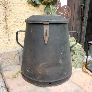 Könnte beinhalten: Ein schwarzer Metallbehälter mit einem Scharnierdeckel und zwei Griffen. Der Behälter hat ein abgenutztes, rustikales Aussehen und ist aus Metall.